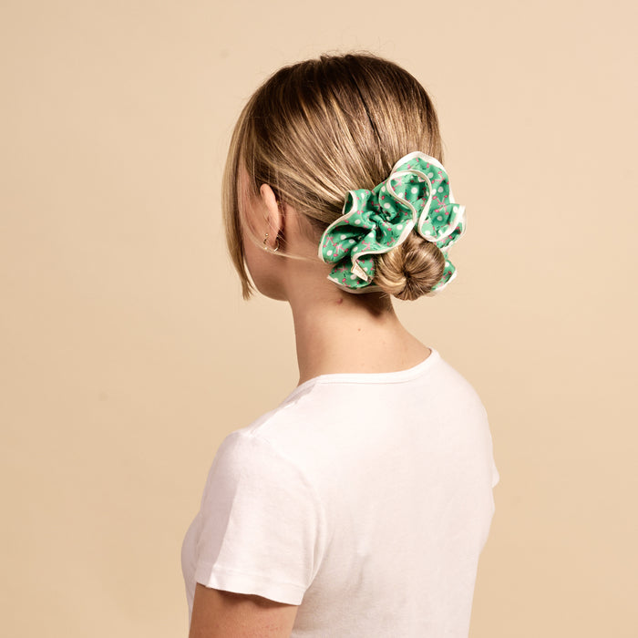 A blonde woman with her hair in a bun wears The Darling Effect's Tee Time Ruffle Up Scrunchie, a large green polka-dot ruffled accessory.