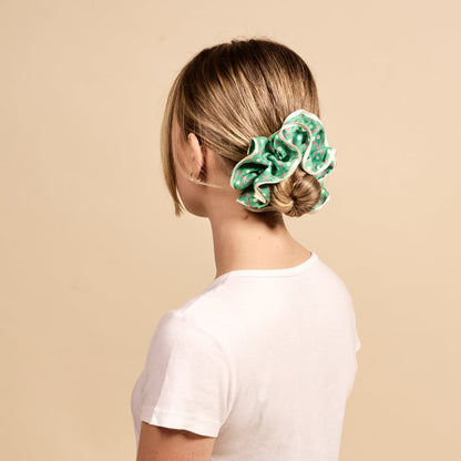 A blonde woman with her hair in a bun wears The Darling Effect's Tee Time Ruffle Up Scrunchie, a large green polka-dot ruffled accessory.