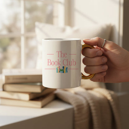 A hand holds The Darling Effect’s The Book Club 11 oz Cozy Cute Ceramic Mug, featuring pink lettering above a vibrant bookshelf. Ideal for coffee or tea, the golden handle and soft background add charming detail.