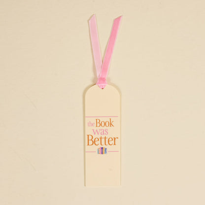 The Darling Effect’s "The Book Was Better" acrylic bookmark features cream coloring, a pink ribbon, pink and gold text, and a cute illustration of three colorful books at the bottom.