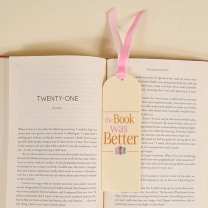 An open book displays chapter twenty-one, topped with The Book Was Better Acrylic Bookmark by The Darling Effect—a cream acrylic piece with a pink ribbon and “The Book Was Better” in pink and orange text—perfect for book lovers.