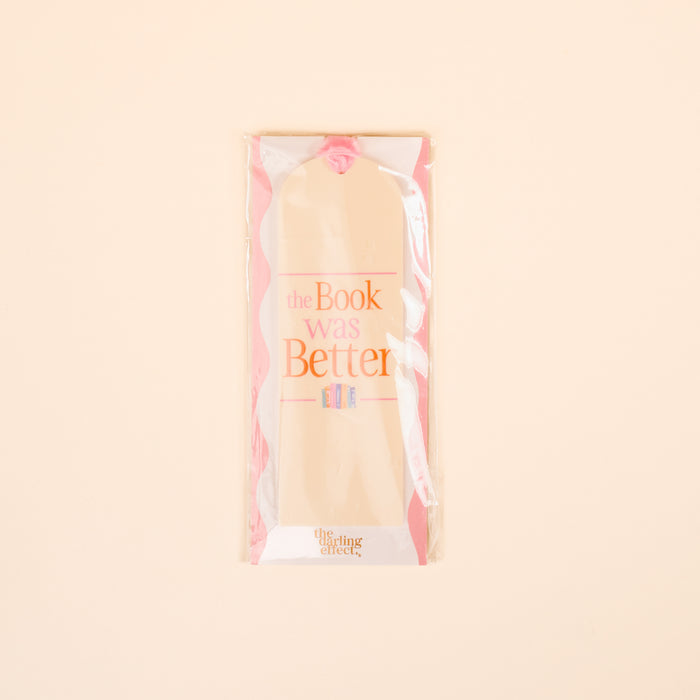 The Darling Effect’s “The Book Was Better” Acrylic Bookmark features beige with pink edges and pink-orange text. Packaged in clear plastic on a pale peach background, it’s an ideal gift for readers who love unique bookmarks.