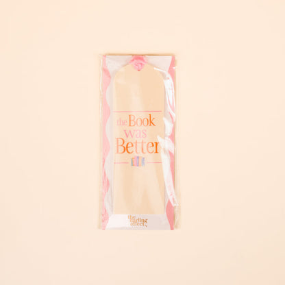 The Darling Effect’s “The Book Was Better” Acrylic Bookmark features beige with pink edges and pink-orange text. Packaged in clear plastic on a pale peach background, it’s an ideal gift for readers who love unique bookmarks.