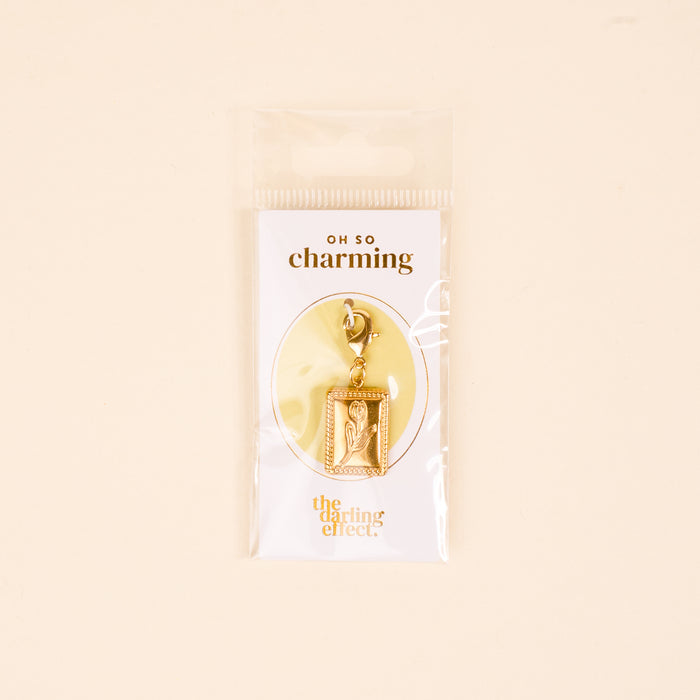 Add unique flair to your charm jewelry with the Tulip Time Jewelry Charm Gold by The Darling Effect—a gold-tone tulip in a rectangular frame on a lobster clasp. Comes in plastic packaging with “OH SO charming” and The Darling Effect branding.