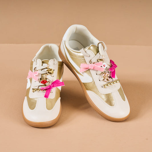 Two stylish sneakers with white and gold panels, adorned with pink ribbons, gold chains, and Velvet Crush Dazzle Shoe Charms by The Darling Effect for a playful Valentine’s look, displayed on a beige surface.