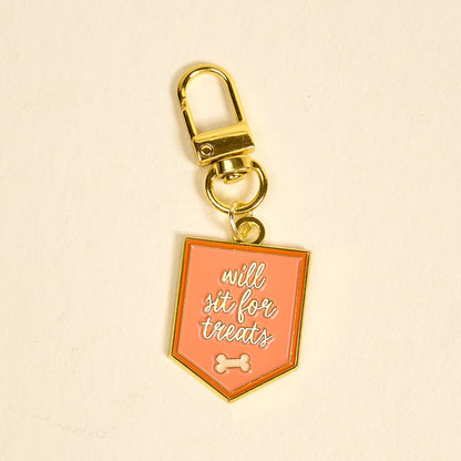 The Darling Effect’s Will Sit for Treats Pawfect Collar Charm features a gold keychain with a pink shield tag, white script, and bone icon—perfect for adding playful style to your pet accessories collection.