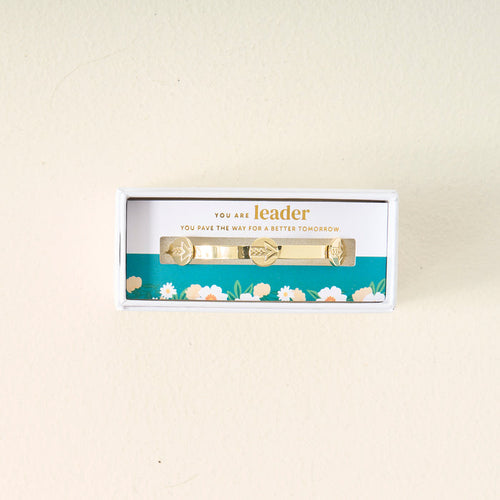 The Darling Effect's "You Are A Leader" Skinny Stacker Trait Bracelet features 14k gold plating and leaf charms, presented in a blue floral box with “You are leader. You pave the way for a better tomorrow” on the insert.