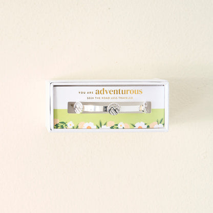 Gift-ready and boxed, The Darling Effect's You Are Adventurous Skinny Stacker Bracelet features three charms and includes a card that reads, “You are adventurous. Seek the road less travelled,” with a floral design below.