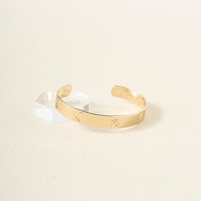 The Darling Effect’s “You Are Ambitious” cuff bracelet, engraved with meaningful symbols, rests on a light textured surface beside a clear rectangular block—a thoughtful and beautiful gift.