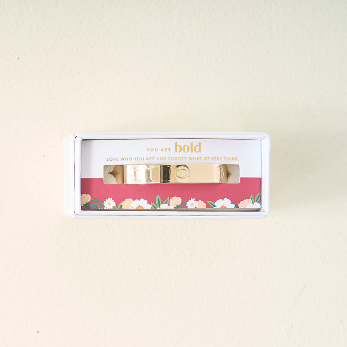 The Darling Effect’s You Are Bold Cuff Bracelet, engraved with “You are bold. Love who you are and forget what others think.,” is displayed in a floral white box on a cream backdrop—a meaningful gift for any occasion.