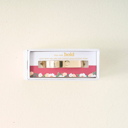 The Darling Effect’s You Are Bold Cuff Bracelet, engraved with “You are bold. Love who you are and forget what others think.,” is displayed in a floral white box on a cream backdrop—a meaningful gift for any occasion.