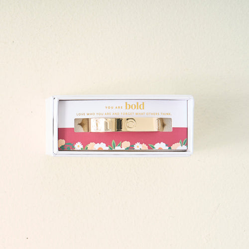 The Darling Effect’s You Are Bold Cuff Bracelet, engraved with “You are bold. Love who you are and forget what others think.,” is displayed in a floral white box on a cream backdrop—a meaningful gift for any occasion.