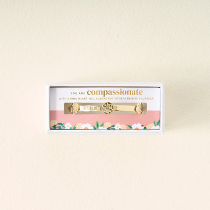 The Darling Effect You Are Compassionate Skinny Stacker Bracelet, a 14k gold plated adjustable cuff with floral designs, is displayed in a box featuring a floral border and card that reads: “You are compassionate. With a kind heart you always put others before yourself.”.