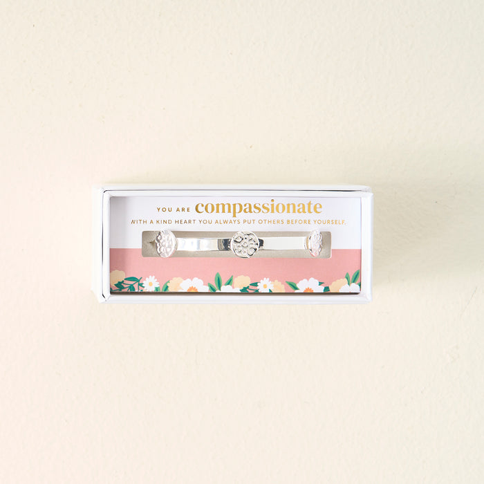 The Darling Effect’s You Are Compassionate Skinny Stacker Bracelet—a silver adjustable cuff with heart designs—comes gift-ready in a floral box featuring the message: “You are compassionate. With a kind heart you always put others before yourself.”.