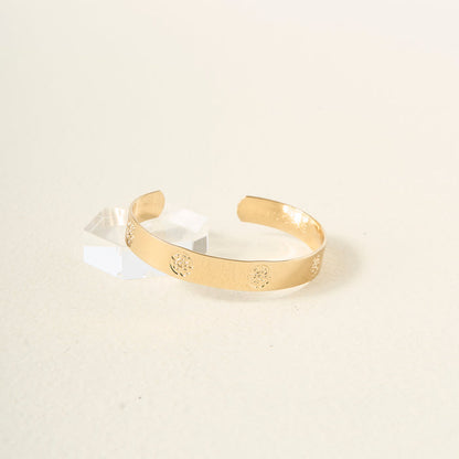 The Darling Effect's "You Are Compassionate Trait Cuff Bracelet," a gold cuff engraved with floral patterns, is displayed on a light surface beside a clear geometric block, embodying timeless elegance.