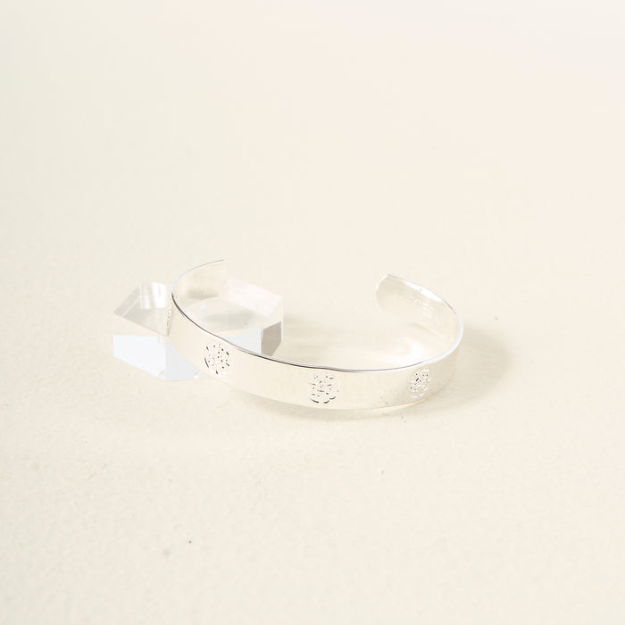 The You Are Compassionate Trait Cuff Bracelet by The Darling Effect, a shiny sterling silver piece with engraved floral details, is displayed near a clear geometric acrylic block on a light cream background.