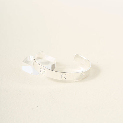 The You Are Compassionate Trait Cuff Bracelet by The Darling Effect, a shiny sterling silver piece with engraved floral details, is displayed near a clear geometric acrylic block on a light cream background.
