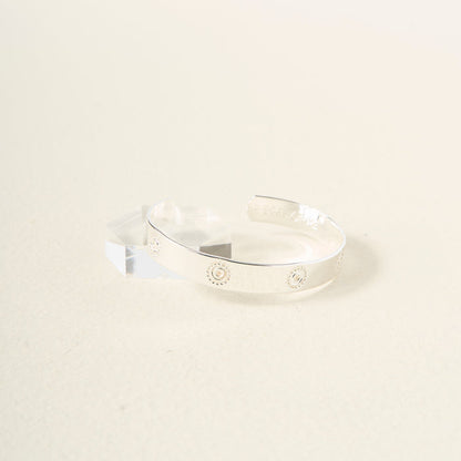 The Darling Effect's You Are Confident Trait Cuff Bracelet, crafted in sterling silver with engraved sunburst designs, rests on a light textured surface beside a clear geometric block—making it a meaningful gift for any occasion.