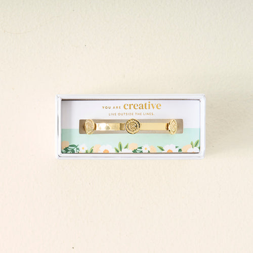 The Darling Effect’s You Are Creative Skinny Stacker Bracelet is presented in a white floral box with a jewelry card that reads, "You are creative. Live outside the lines.