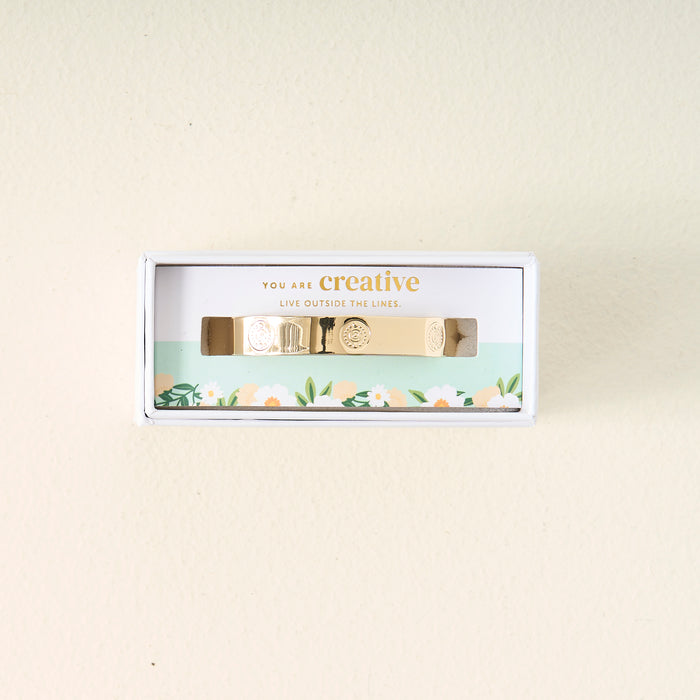 The Darling Effect’s You Are Creative Trait Cuff Bracelet, featuring engraved designs, is shown in a floral white box with the message “You are creative. Live outside the lines,” making it a thoughtful gift on a light beige backdrop.