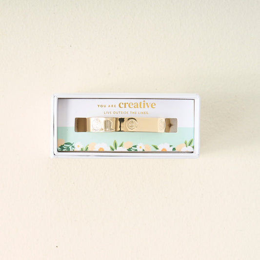 The Darling Effect’s You Are Creative Trait Cuff Bracelet, featuring engraved designs, is shown in a floral white box with the message “You are creative. Live outside the lines,” making it a thoughtful gift on a light beige backdrop.