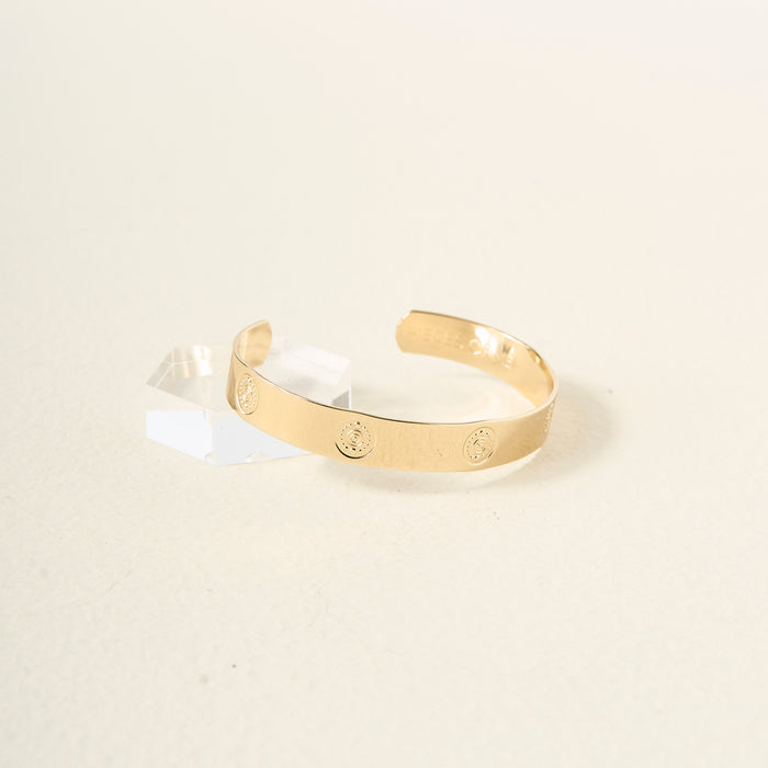 The Darling Effect's "You Are Creative" Trait Cuff Bracelet features a smooth gold finish with small circular engravings, beautifully displayed on a light surface beside a clear acrylic block—an elegant and meaningful gift for any occasion.