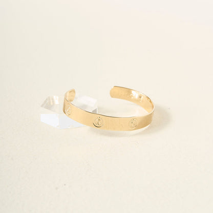 The Darling Effect's "You Are Creative" Trait Cuff Bracelet features a smooth gold finish with small circular engravings, beautifully displayed on a light surface beside a clear acrylic block—an elegant and meaningful gift for any occasion.