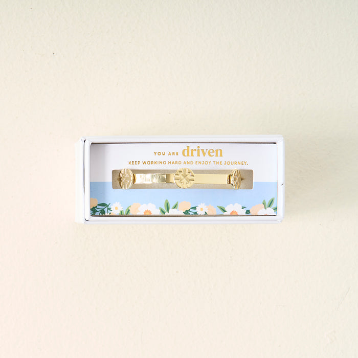 The Darling Effect’s You Are Driven Skinny Stacker Bracelet features sun and leaf designs on 14k gold plating in a white gift box. A blue sky and flower border frames “You are driven. Keep working hard and enjoy the journey.”.