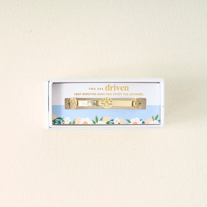 The Darling Effect’s You Are Driven Skinny Stacker Bracelet features sun and leaf designs on 14k gold plating in a white gift box. A blue sky and flower border frames “You are driven. Keep working hard and enjoy the journey.”.