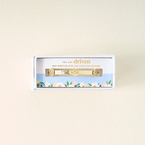 The Darling Effect’s You Are Driven Skinny Stacker Bracelet features sun and leaf designs on 14k gold plating in a white gift box. A blue sky and flower border frames “You are driven. Keep working hard and enjoy the journey.”.