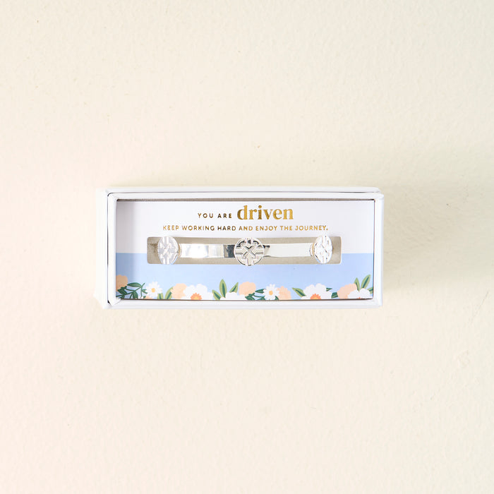 The Darling Effect’s You Are Driven Skinny Stacker Bracelet is a 14k gold plated adjustable cuff with engraved symbols, gift boxed with the message "You are driven. Keep working hard and enjoy the journey" and a floral accent.