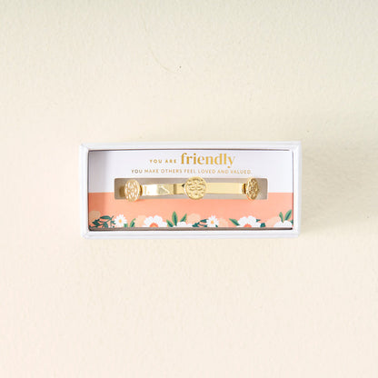 The Darling Effect You Are Friendly Skinny Stacker Bracelet, a 14k gold plated adjustable cuff, comes in a white floral box with a card that says, "You are friendly. You make others feel loved and valued." Perfect gift-ready jewelry.