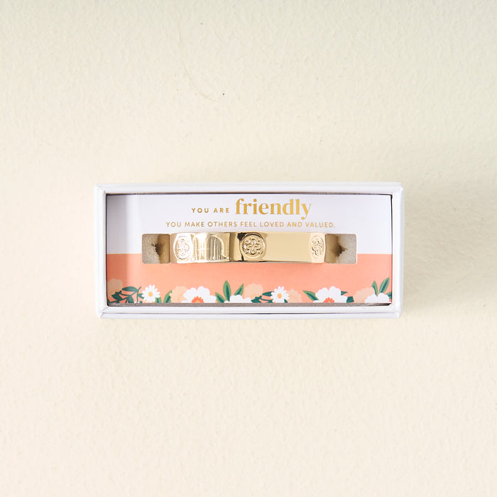 The Darling Effect’s You Are Friendly Trait Cuff Bracelet is displayed in a white box with floral edging. Inside, a card reads: YOU ARE friendly. YOU MAKE OTHERS FEEL LOVED AND VALUED—a meaningful gift with engraved designs.