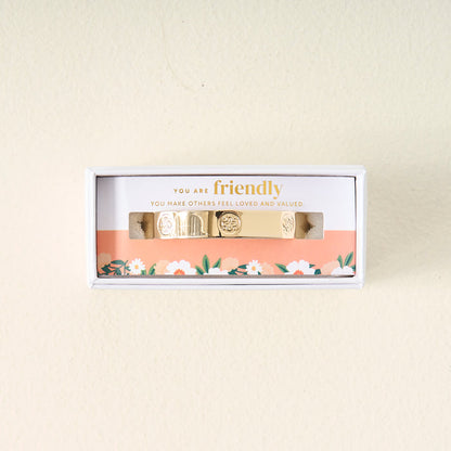 The Darling Effect’s You Are Friendly Trait Cuff Bracelet is displayed in a white box with floral edging. Inside, a card reads: YOU ARE friendly. YOU MAKE OTHERS FEEL LOVED AND VALUED—a meaningful gift with engraved designs.