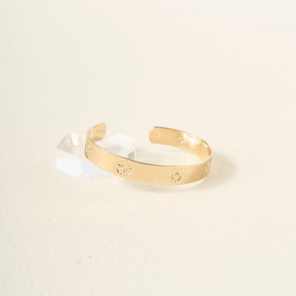 The You Are Loyal Cuff Bracelet by The Darling Effect, featuring subtle engraved patterns in gold, is displayed beside a small, clear geometric crystal on a light textured surface.