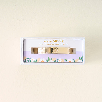 The Darling Effect's You Are Sassy Cuff Bracelet, featuring engraved designs, comes in a white box with a floral border. The packaging says "YOU ARE SASSY" and "KEEP IT CLASSY AND ALWAYS SASSY.
