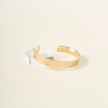 The You Are Sassy Cuff Bracelet by The Darling Effect, featuring engraved circular designs and an adjustable gold band, rests on a light textured surface beside a small clear geometric block.