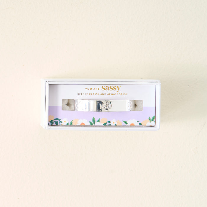 A small box from The Darling Effect holds the adjustable You Are Sassy Cuff Bracelet in sterling silver. The packaging reads "YOU ARE sassy. KEEP IT CLASSY AND ALWAYS SASSY" above a floral border on a light cream background.