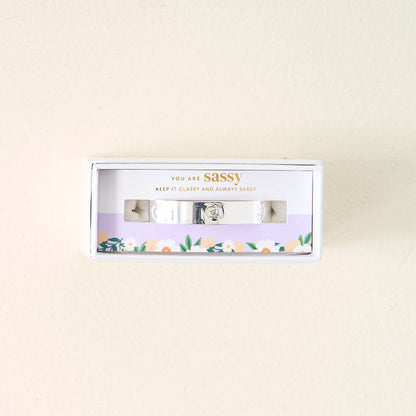 A small box from The Darling Effect holds the adjustable You Are Sassy Cuff Bracelet in sterling silver. The packaging reads "YOU ARE sassy. KEEP IT CLASSY AND ALWAYS SASSY" above a floral border on a light cream background.